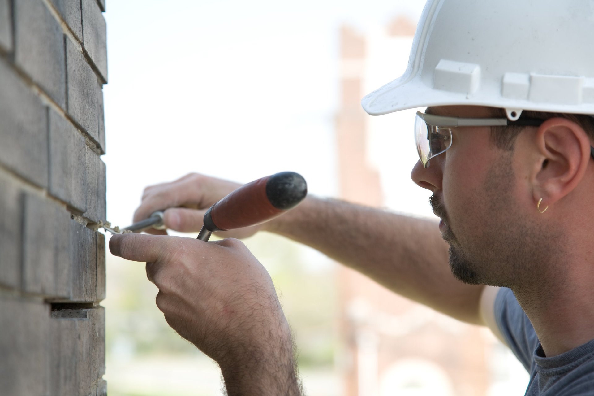 A construction worker wearing a hard hat and safety glasses uses a tool to work on a brick wall