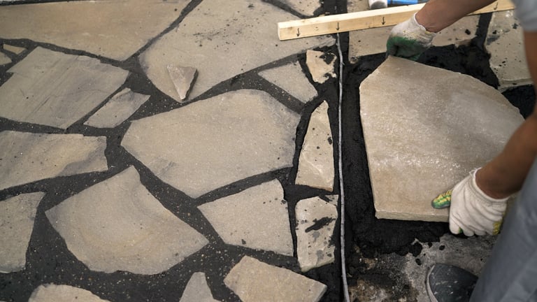 Construction worker carefully placing irregular flagstone on a bed of dark mortar, creating a decorative pathway