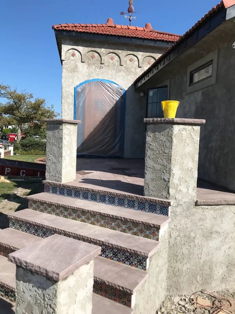 Stone church entrance with decorative arch, red-tiled roof, concrete steps, and blue door under clear sky
