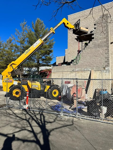 Yellow telehandler lifting a steel frame attachment against a brick building on a construction site
