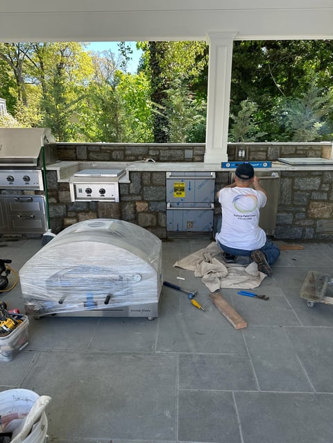 Worker installing outdoor kitchen appliances on a stone patio with wrapped equipment and tools visible