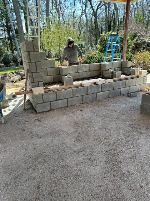 Man building outdoor concrete block structure in wooded yard with blue ladder and construction materials