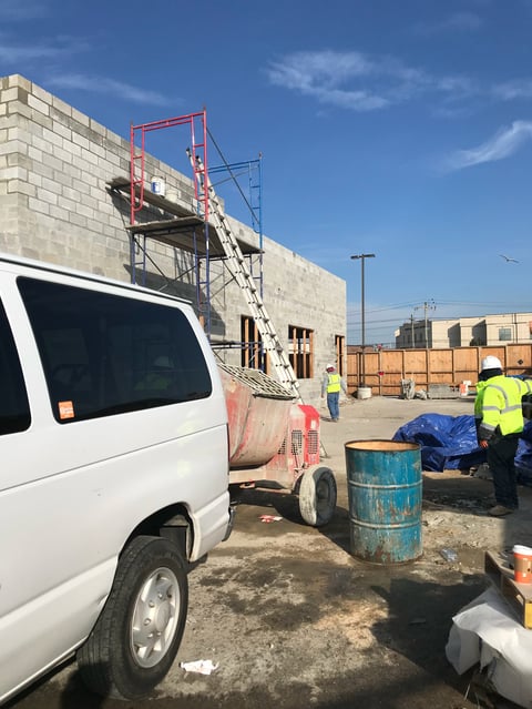 Construction site with white van, red metal scaffolding on concrete building, workers in safety vests, and blue barrels under clear sky