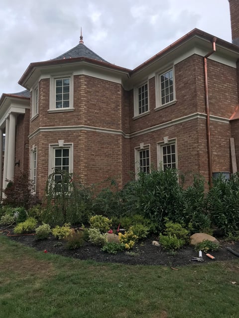 Two-story brick house with turret, multiple windows, landscaped front yard with shrubs and lawn under cloudy sky