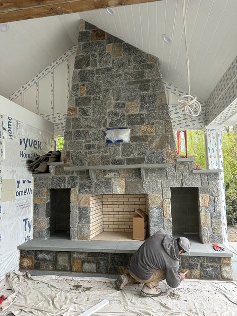 Worker in protective gear constructing a large stone fireplace with brick interior in an unfinished room