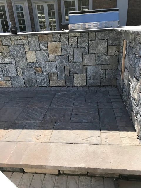 Outdoor patio with geometric stone paving in varying shades of gray and beige, featuring a modern glass building and blue signage in the background