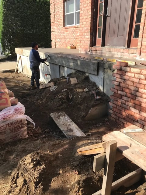 Construction worker installing concrete foundation against red brick house with exposed excavation and materials