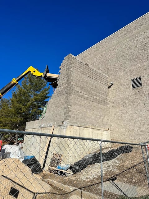 Yellow construction crane lifting equipment near brick building under clear blue sky with chain-link fence in foreground