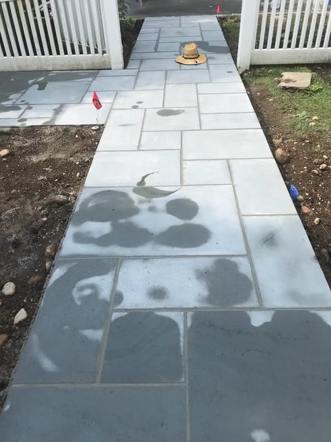 Concrete patio pathway with shadow patterns and paving stones, flanked by white fences and landscaping