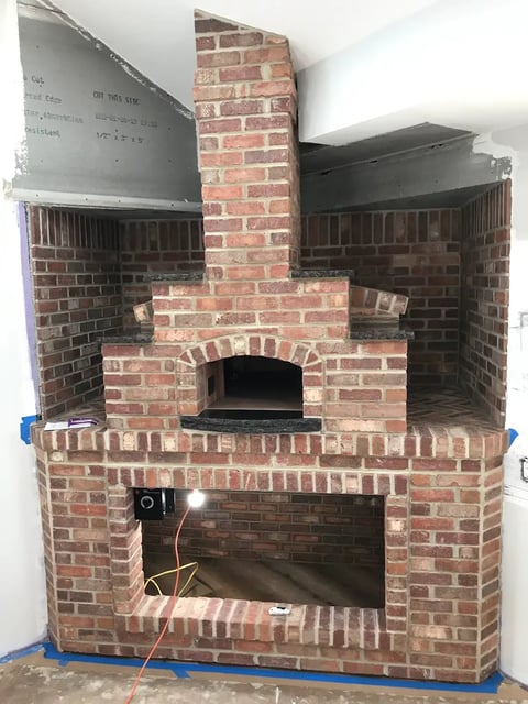 Red brick fireplace and pizza oven under construction with metal ductwork and white walls