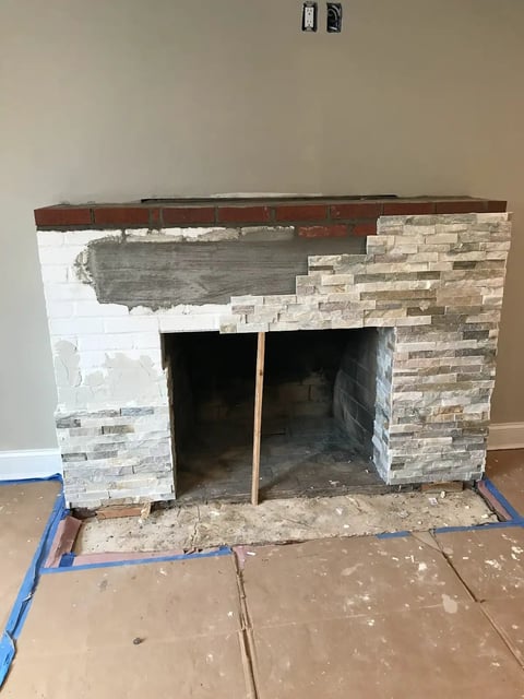 Fireplace under renovation with exposed brick, stone veneer, and white painted surfaces, surrounded by protective blue tape on floor