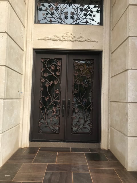 Ornate black double doors with floral ironwork design in a cream stone entryway with transom window above