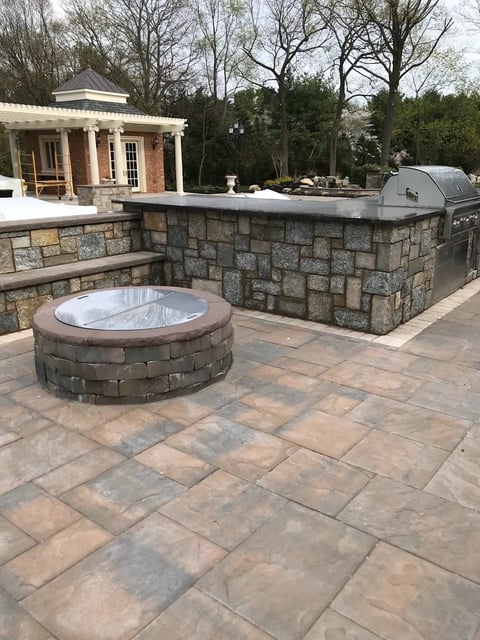 Backyard patio with stone fire pit, outdoor kitchen with grill, and pool house in background