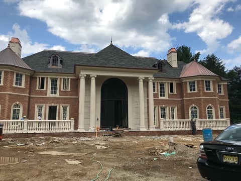 Large brick mansion under construction with white columns and portico, set against cloudy blue sky with construction materials visible on grounds