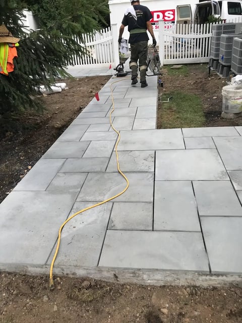 A yellow extension cord running across a newly laid gray stone paver walkway with a worker in dark clothing visible in the background