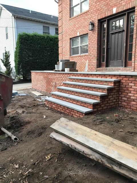Brick house entrance under construction with new concrete steps, exposed soil, and building materials scattered around the foundation area