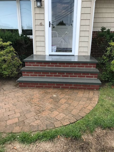 White house entrance with brick steps and dark top landing, surrounded by hedges on a brick paver patio