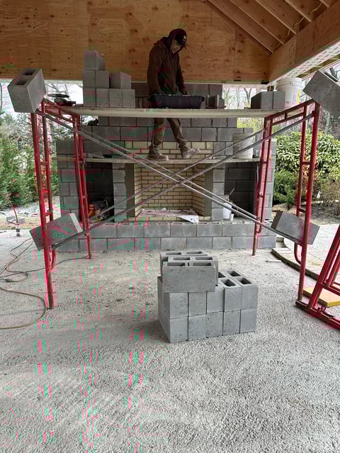 Worker constructing a brick fireplace structure surrounded by red metal scaffolding on a construction site