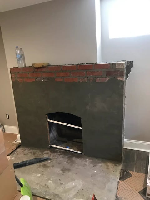 Fireplace under construction with exposed brick top, dark metal surround, and concrete base in a residential room